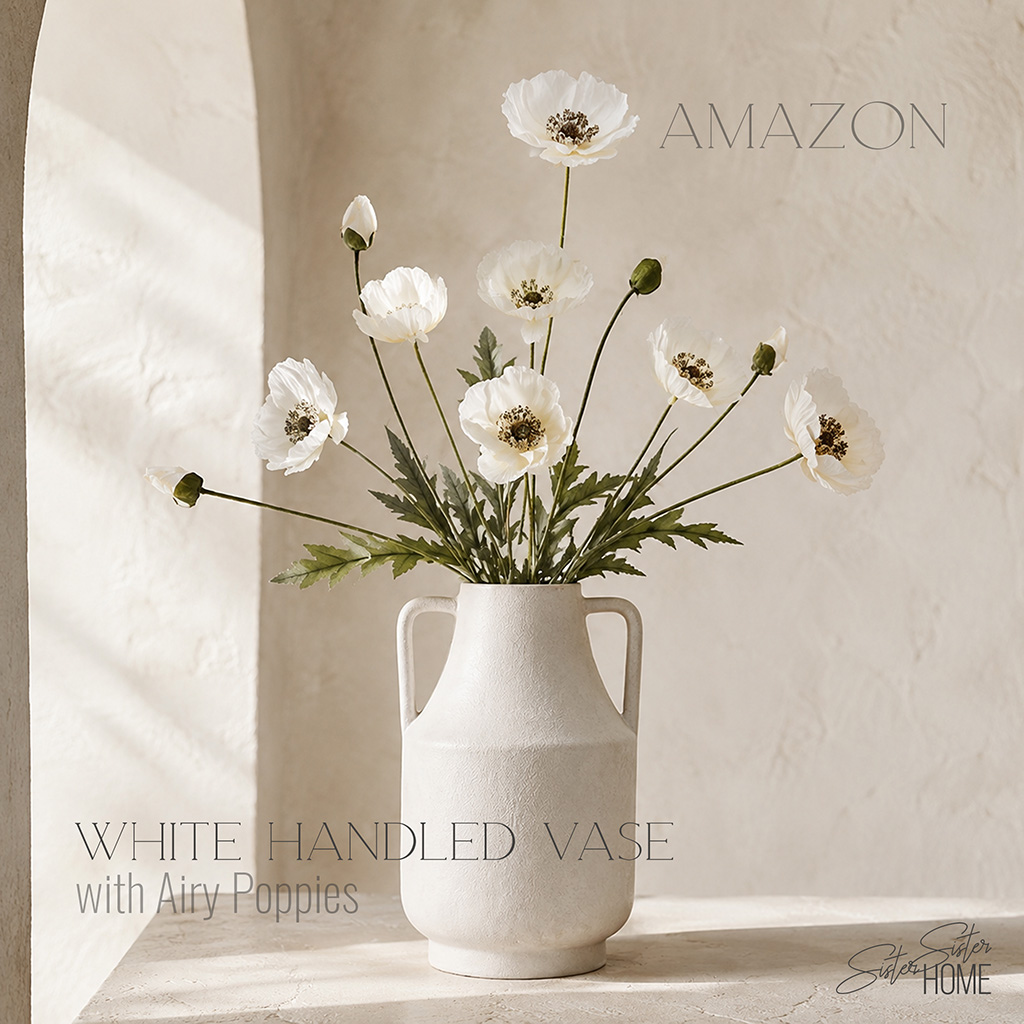 White ceramic handled vase filled with airy white poppies in a sunlit Mediterranean-inspired setting.